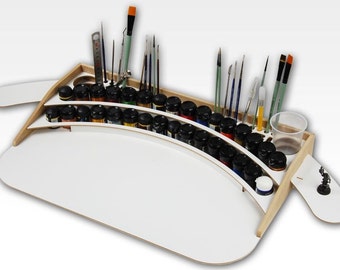 May include: A white wooden paint station with a curved surface for holding paint bottles. The station has a built-in brush holder and a small cup for water. There are 24 paint bottles in the station, and several brushes are in the holder.