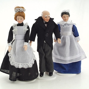 Puede incluir: Tres muñecas de porcelana vestidas con atuendos de sirvienta y mayordomo de estilo vintage. Las muñecas femeninas llevan delantales blancos y vestidos negros con ribetes de encaje. La muñeca masculina lleva un traje negro con camisa blanca y corbata de lazo.