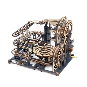 May include: A mechanical marble run constructed from laser-cut wood pieces in natural and dark gray tones. The structure features a complex system of gears, a hand crank, tracks, and a central spiral funnel designed for small metal ball bearings.