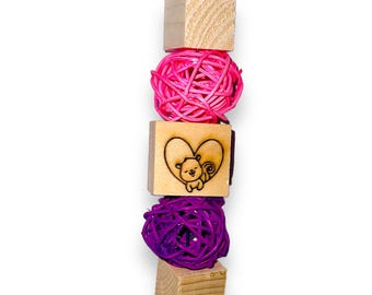 May include: A wooden toy for small animals, featuring a pink and purple woven ball, a wooden block with a heart and a squirrel design, and a wooden block with a heart design.