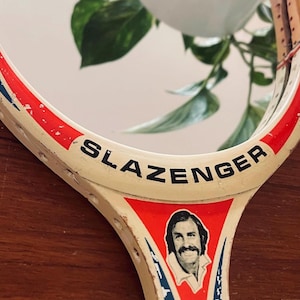 Op de afbeelding: Een vintage Slazenger tennisracket met een rood, wit en blauw ontwerp. De racket heeft aan de ene kant een spiegel en aan de andere kant een foto van een man.