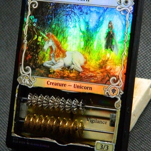 May include: A framed "Lonesome Unicorn" card with a fantasy illustration of a unicorn in a forest. The card is modified with an abacus, featuring silver and gold beads. The card reads "Creature - Unicorn" and "Vigilance".