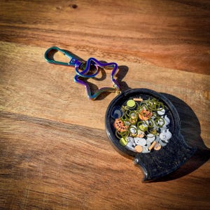 Puede incluir: Un llavero negro con forma de cabeza, lleno de abalorios temáticos de Halloween. El llavero tiene un cierre iridiscente en forma de estrella. En el interior hay calabazas, fantasmas y otras formas festivas.