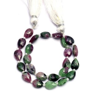 May include: A strand of green and pink gemstone beads with a pear shape. The beads are strung on a white cord with white tassels at the ends.