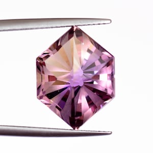 Könnte beinhalten: Ein facettierter, sechseckiger Edelstein, der von einer Pinzette gehalten wird. Der Edelstein weist einen Farbverlauf aus Rosa- und Lilatönen mit einem zentralen Lichtpunkt auf. Der Schliff erzeugt einen Sternausbruch-Effekt und betont die Klarheit des Steins. Der Hintergrund ist weiß.