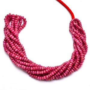 Könnte beinhalten: Eine Kette aus rosafarbenen Rubinperlen, auf einer roten Schnur aufgefädelt. Die Perlen sind rund und haben eine glatte, polierte Oberfläche.
