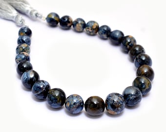May include: A strand of polished, round Pietersite gemstone beads. The beads are primarily dark blue with swirling patterns of gold and brown. A silver tassel is attached to the end of the strand.