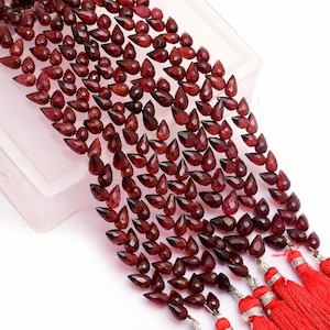 May include: A strand of deep red gemstone beads in a teardrop shape. The beads are strung on a silver wire and have a tassel at the end.