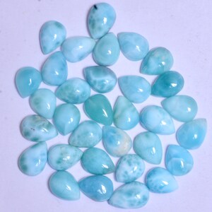 May include: A collection of 30 pear-shaped, iridescent blue and white gemstone cabochons. The stones have a smooth, polished finish and are arranged in a loose circle on a white background.