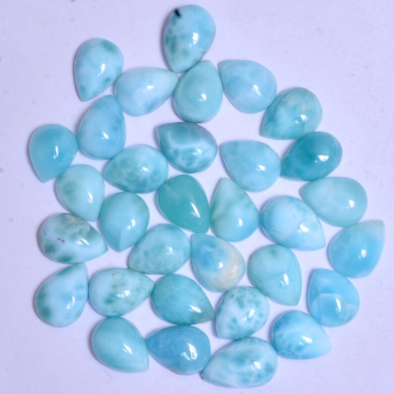 May include: A collection of 30 pear-shaped, iridescent blue and white gemstone cabochons. The stones have a smooth, polished finish and are arranged in a loose circle on a white background.
