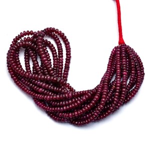Op de afbeelding: Een streng dieprode robijn kralen, geregen aan een rood koord. De kralen zijn rond en glad, en ze zijn gerangschikt in een los, vloeiend patroon.