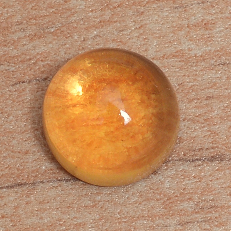 May include: A round, orange gemstone with a smooth, polished surface. The stone has a translucent appearance with a slight shimmer.