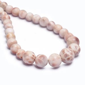 Puede incluir: Una hebra de cuentas de concha de color rosa pálido con una superficie texturizada. Las cuentas son redondas y tienen un aspecto natural y orgánico.