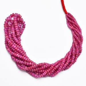 Könnte beinhalten: Eine Strähne aus rosafarbenen Rubinperlen, auf einem roten Faden aufgefädelt. Die Perlen sind klein und rund, mit einer glatten, polierten Oberfläche.