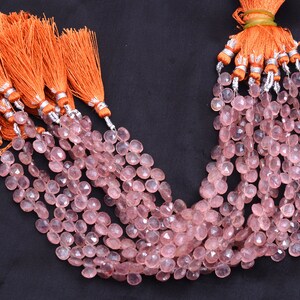 May include: Pink gemstone beads in a teardrop shape, strung together and topped with orange tassels with silver detailing. The beads are displayed against a dark background. Each strand is a collection of beads.