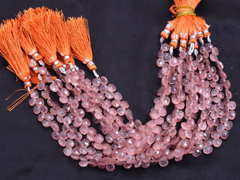 May include: Pink gemstone beads in a teardrop shape, strung together and topped with orange tassels with silver detailing. The beads are displayed against a dark background. Each strand is a collection of beads.