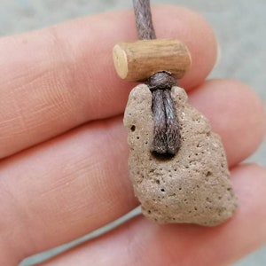 Könnte beinhalten: Ein brauner Steinanhänger mit rauer Textur, befestigt an einer braunen Schnur mit einer Holzperle. Der Anhänger hat die Form eines Herzens.