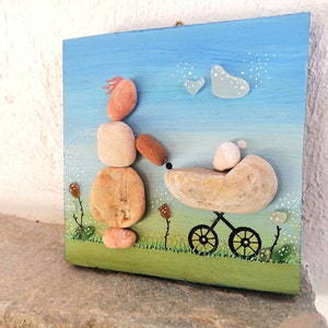 May include: A painted wooden panel with a pebble art design depicting a person pushing a baby carriage. The person is made of stacked stones in shades of pink, brown, and beige. The carriage is made of a single large stone with a smaller stone representing the baby. The background is painted blue with white dots and two white hearts.