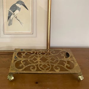 May include: A framed bird print with a gold frame rests on a wooden surface. In front of the print is a decorative brass trivet with ornate cut-out designs and small claw feet. A small bouquet of white flowers is in the background.