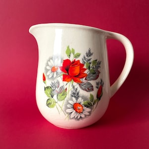 May include: White ceramic jug with a floral design featuring red roses and white daisies. The jug has a textured surface and a handle.