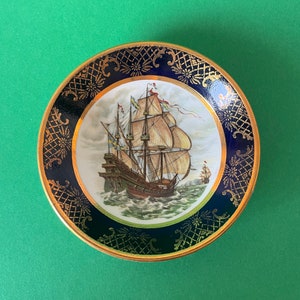 May include: A decorative plate with a gold and blue border features a painting of a ship with white sails against a blue and white background.