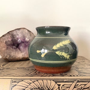 May include: A small, green ceramic vase with a brown rim and a yellow floral design. The vase is sitting on a book with a brown cover.