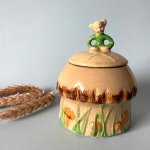 May include: A ceramic sugar bowl shaped like a mushroom, with a green and brown glaze. The lid features a small figurine of a person sitting on top of the mushroom. The bowl is decorated with brown mushrooms and green grass.