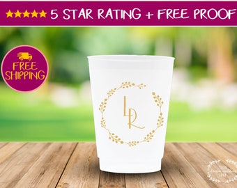 May include: White plastic cup with a gold monogram "LR" surrounded by a wreath of gold leaves. The cup is sitting on a wooden surface.