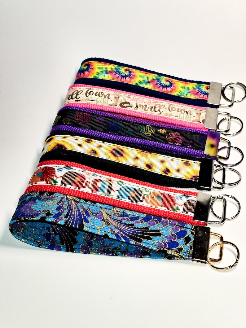 May include: A stack of colourful keychains with different patterns and designs. The keychains feature tie-dye, sunflowers, elephants, and a floral pattern. Each keychain has a gold metal ring.