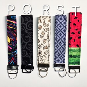 May include: Five colourful keychains with different patterns. The keychains are made of fabric and have a metal ring at the end. The patterns include a swirling abstract design, a black and white swirl pattern, a coffee themed pattern, a blue and white geometric pattern, and a watermelon pattern.