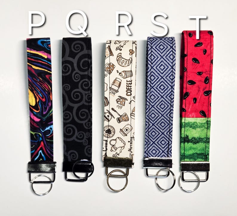 May include: Five colourful keychains with different patterns. The keychains are made of fabric and have a metal ring at the end. The patterns include a swirling abstract design, a black and white swirl pattern, a coffee themed pattern, a blue and white geometric pattern, and a watermelon pattern.