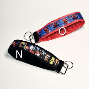 May include: Two keychains with black fabric straps and colourful printed ribbon. One keychain has a red strap with a blue ribbon featuring Spiderman characters. The other keychain has a black strap with a multi-coloured ribbon featuring Star Wars characters.