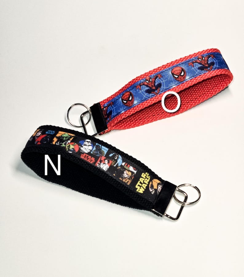May include: Two keychains with black fabric straps and colourful printed ribbon. One keychain has a red strap with a blue ribbon featuring Spiderman characters. The other keychain has a black strap with a multi-coloured ribbon featuring Star Wars characters.