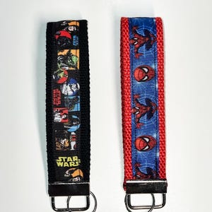 May include: Two keychains with black and red fabric straps. The black strap has a colourful print of Star Wars characters. The red strap has a blue and red print of Spiderman. Both keychains have silver metal rings.