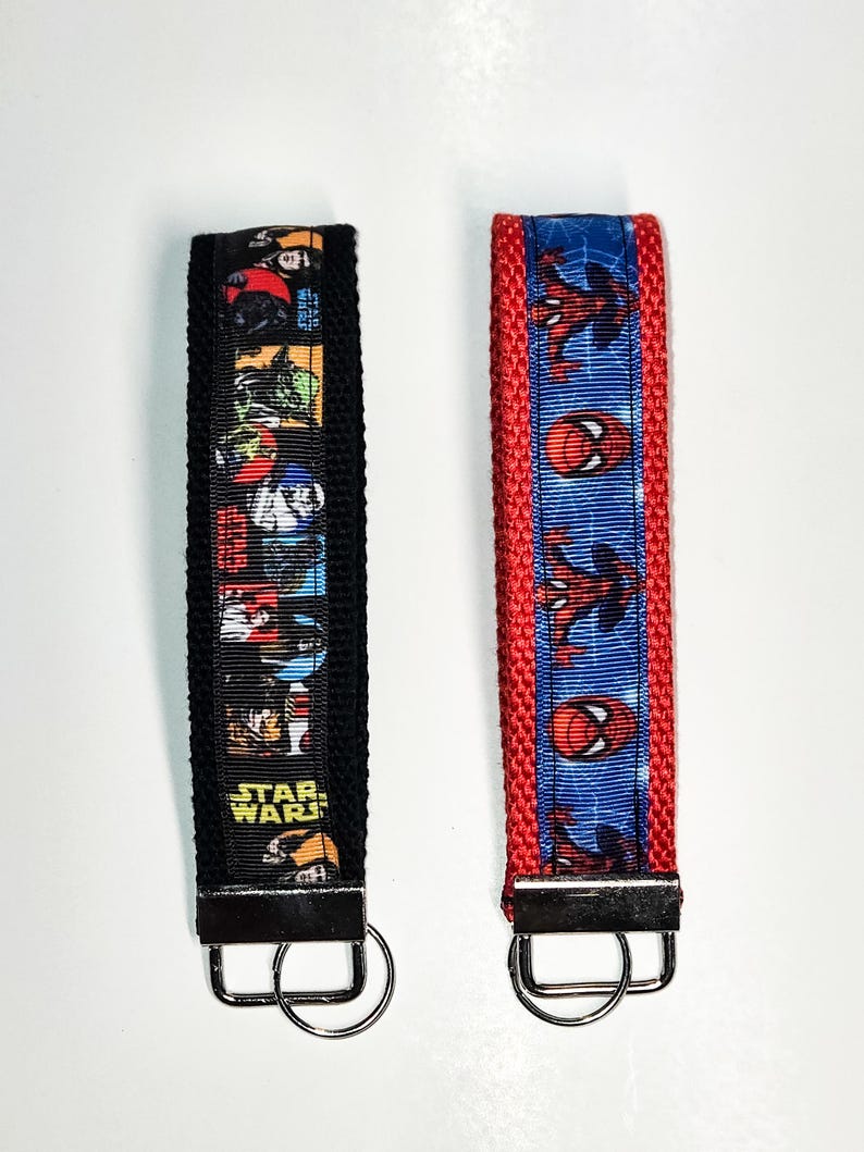 May include: Two keychains with black and red fabric straps. The black strap has a colourful print of Star Wars characters. The red strap has a blue and red print of Spiderman. Both keychains have silver metal rings.