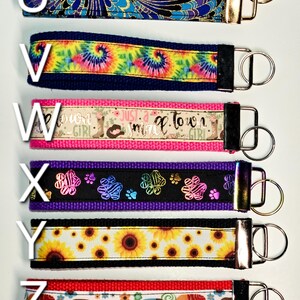 May include: Six colourful keychains with different patterns and designs. The keychains are made of fabric and have a metal ring. The patterns include a blue and gold floral design, a tie-dye pattern, a pink and white pattern with the text "Just a small town girl", a black and purple pattern with paw prints, a yellow and black pattern with sunflowers, and a red pattern with elephants.