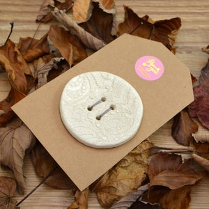 May include: A white ceramic button with a textured surface and two holes. The button is on a brown card with a pink circle sticker that says "Thank You".