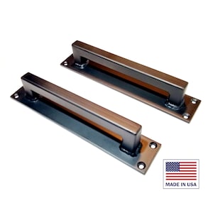 May include: Two black metal door pulls with rectangular plates. The pulls have a square shape and are attached to the plates with screws. Made in the USA.