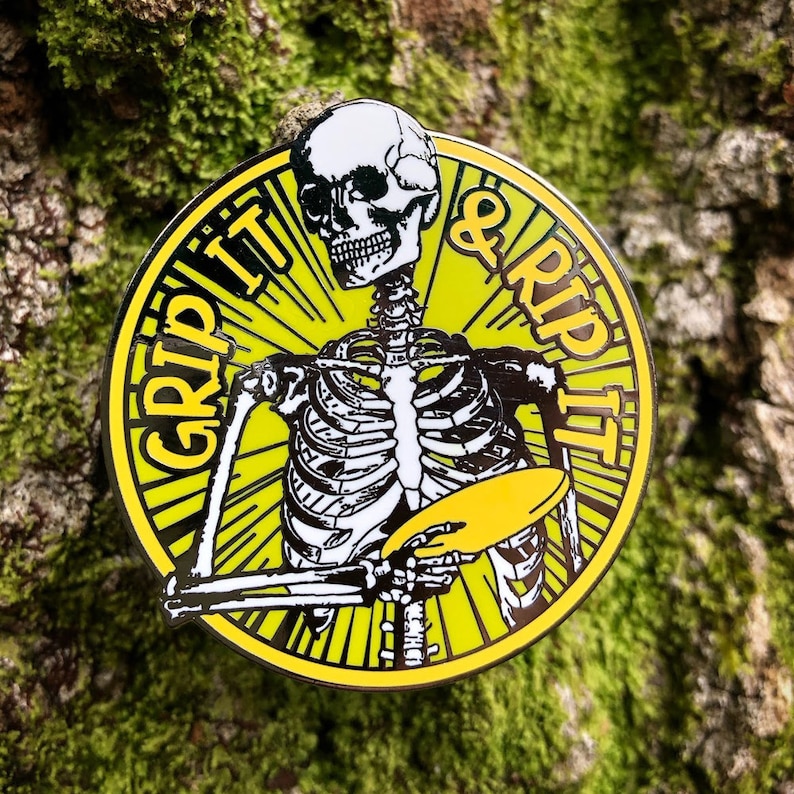 May include: A yellow and black enamel pin featuring a skeleton holding a disc with the text "GRIP IT & RIP IT" around the edge.