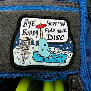 May include: A gray and blue patch with a cartoon narwhal holding a red disc. The text on the patch reads "Bye Buddy Hope you find your DISC".