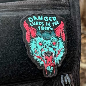 May include: A patch featuring a stylized bat with the text "DANGER LURKS IN THE TREES." The bat has teal fur, red accents, and a long red tongue. The patch is on a black background.