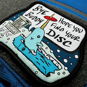 May include: A blue and white embroidered patch with a cartoon narwhal holding a disc golf basket. The text on the patch reads "Bye Buddy Hope You Find Your Disc".