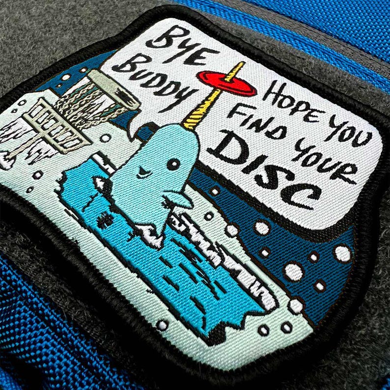 May include: A blue and white embroidered patch with a cartoon narwhal holding a disc golf basket. The text on the patch reads "Bye Buddy Hope You Find Your Disc".