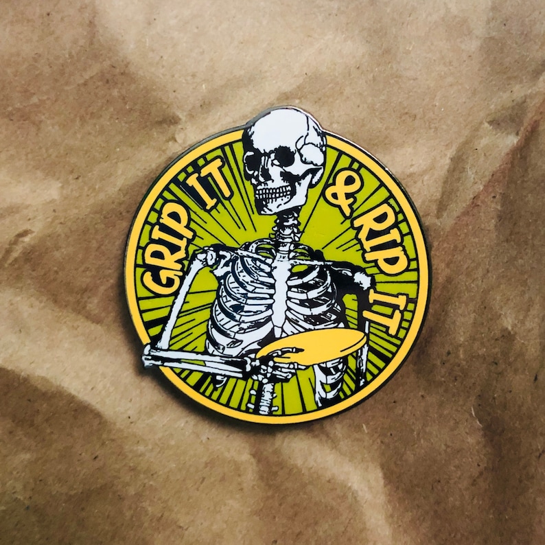 May include: A yellow and green enamel pin featuring a skeleton holding a yellow disc with the text "Grip It & Rip It" in yellow.
