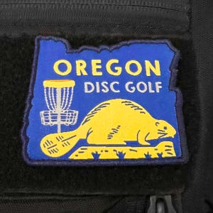 May include: A blue and yellow patch with the text "Oregon Disc Golf" and a cartoon beaver holding a disc golf basket.