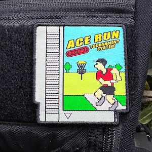 May include: A white and gray patch with a video game-style design. The patch features a blue background with the text "ACE RUN TOURNAMENT SYSTEM" and a pixelated image of a person playing disc golf. The patch has a white border and a small gray triangle at the bottom.