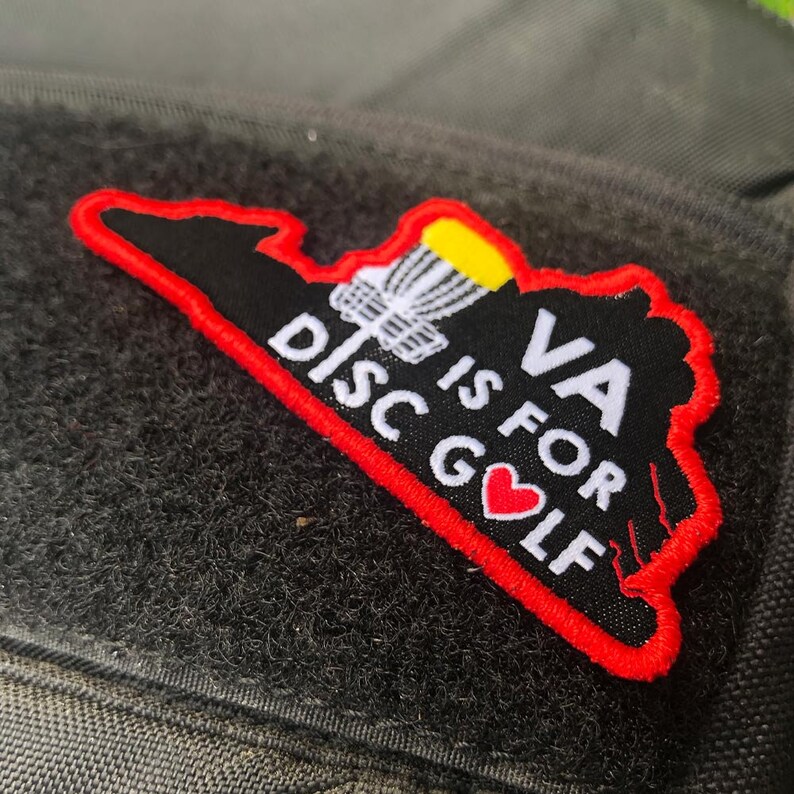 K&ouml;nnte beinhalten: Ein gestickter Patch in Rot, Wei&szlig; und Schwarz, der die Form des Bundesstaates Virginia hat. Der Patch zeigt eine Frisbeescheibe mit gelber Oberseite, den Text "VA is for DISC GOLF" und ein rotes Herz.