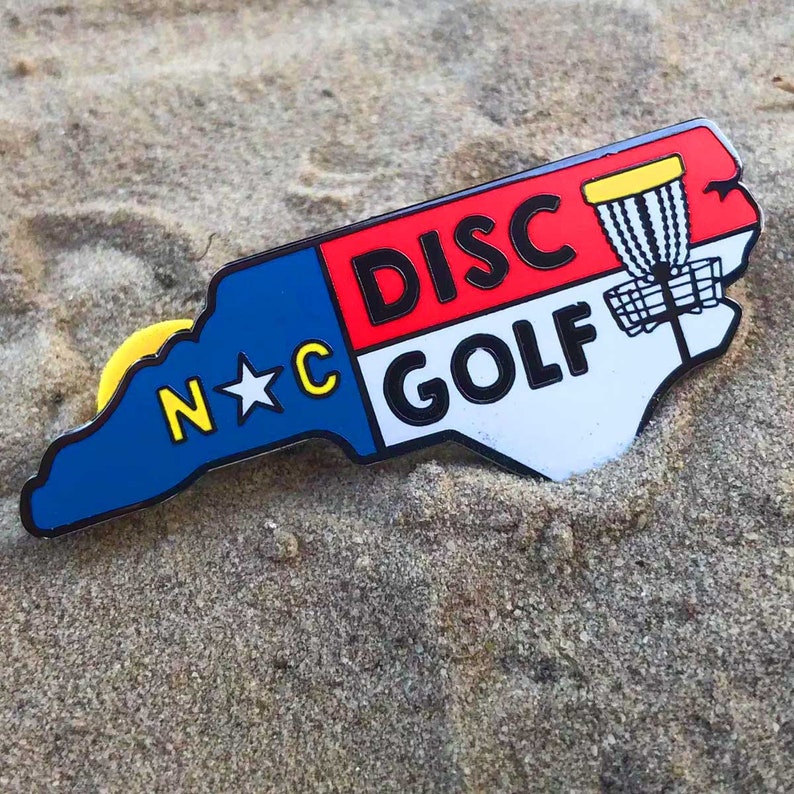 Puede incluir: Un pin de esmalte azul, rojo y blanco con forma del estado de Carolina del Norte. El pin presenta el texto "N*C DISC GOLF" y una canasta de disco golf amarilla.