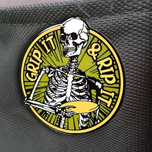 May include: A yellow and green enamel pin featuring a skeleton holding a disc with the text "Grip It & Rip It" in a circular design.