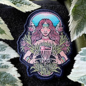 May include: Embroidered patch featuring a woman with long pink hair holding a pink and white cup. The woman is surrounded by green leaves and pink flowers. The patch has a blue background with a light blue sky and white clouds.